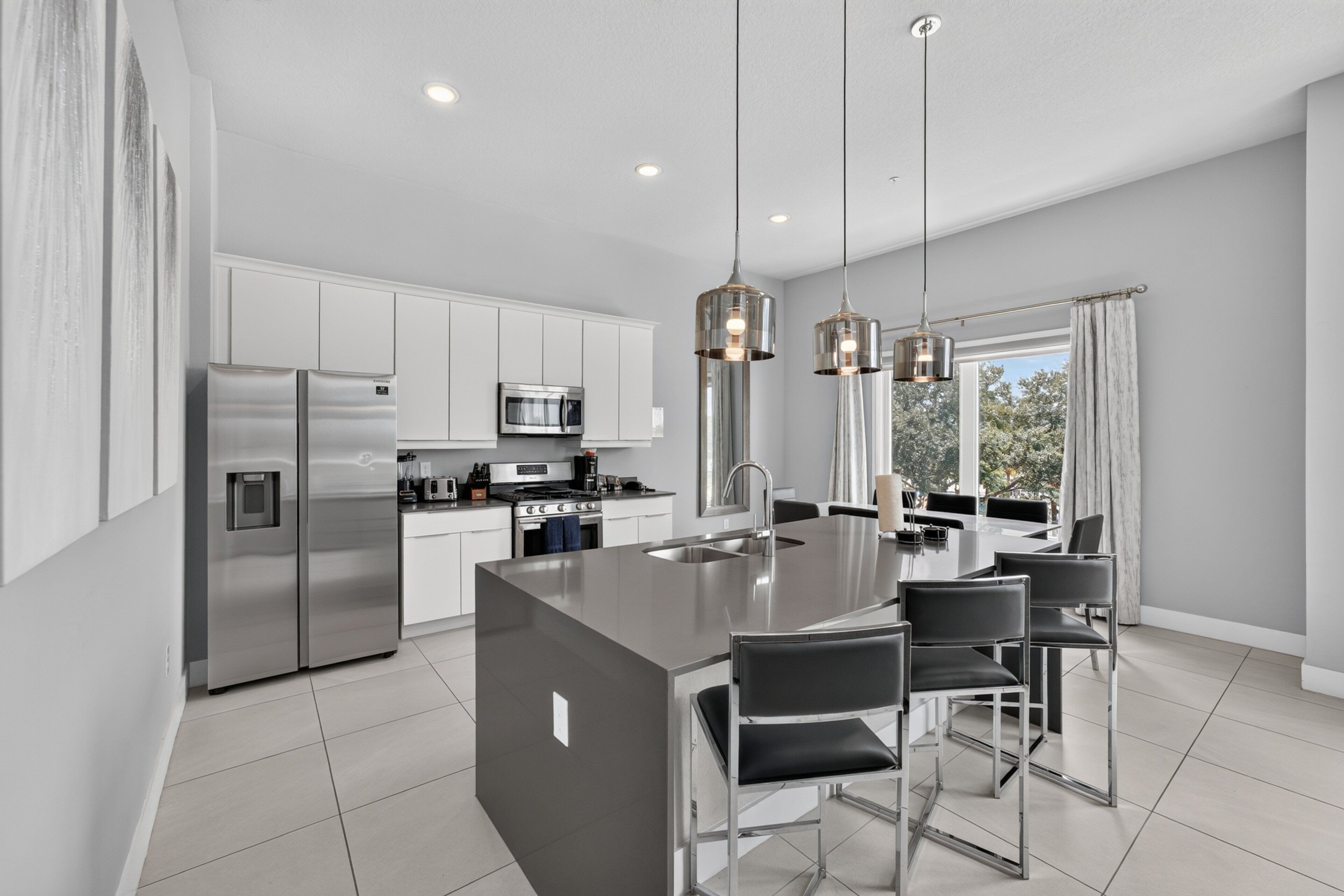 A modern kitchen with stainless steel appliances, a central island with bar stools, pendant lighting, and a view through large windows.