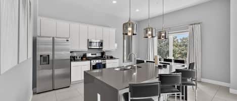 A modern kitchen with stainless steel appliances, a central island with bar stools, pendant lighting, and a view through large windows.