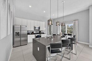 A modern kitchen with stainless steel appliances, a central island with bar stools, pendant lighting, and a view through large windows. - Reunion Resort 5BR Condo | Park View | Resort Pool (Kissimmee)