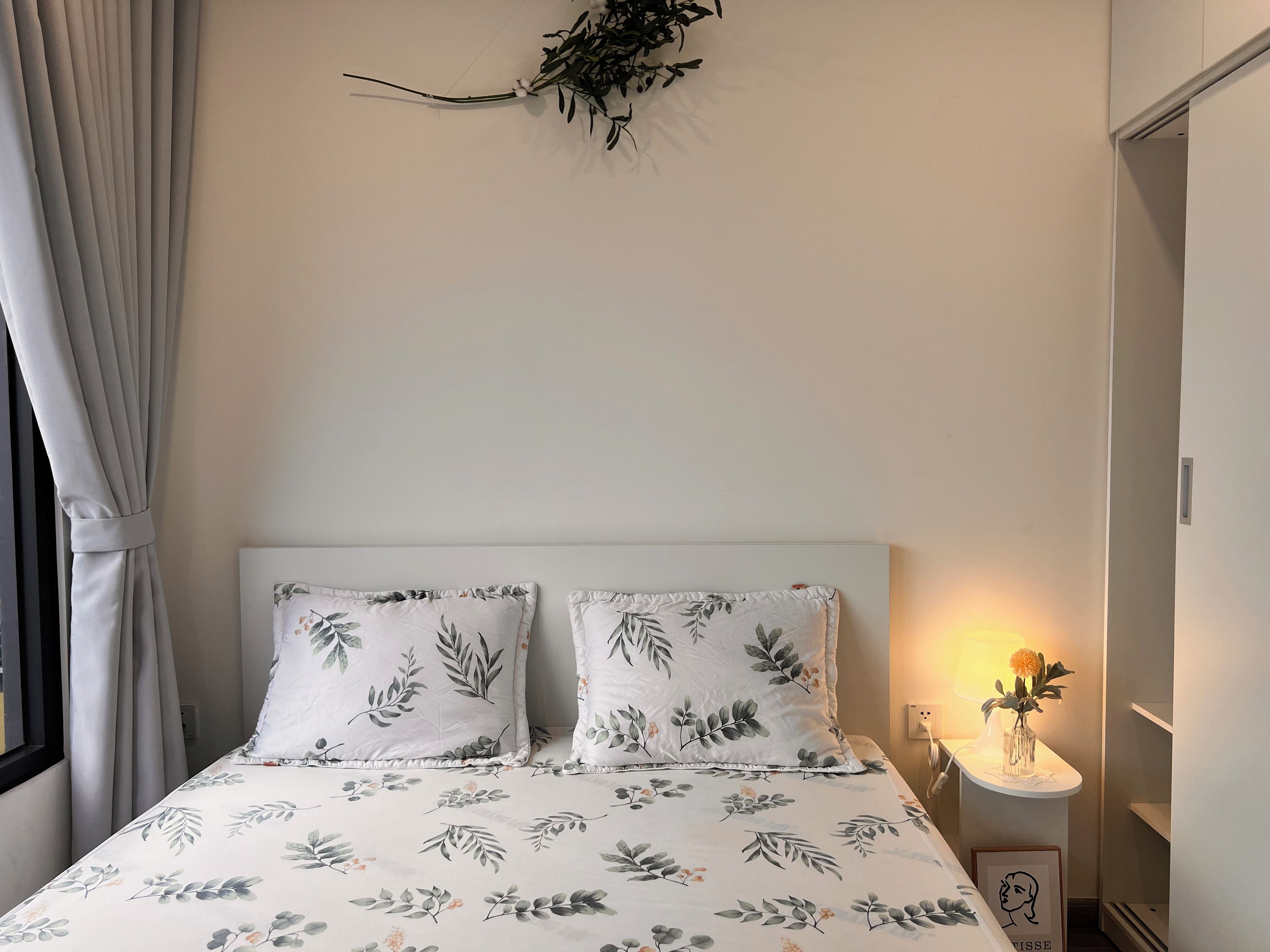 A bedroom with a bed, two pillows, a bedside table with a lamp, and a wall-mounted plant.