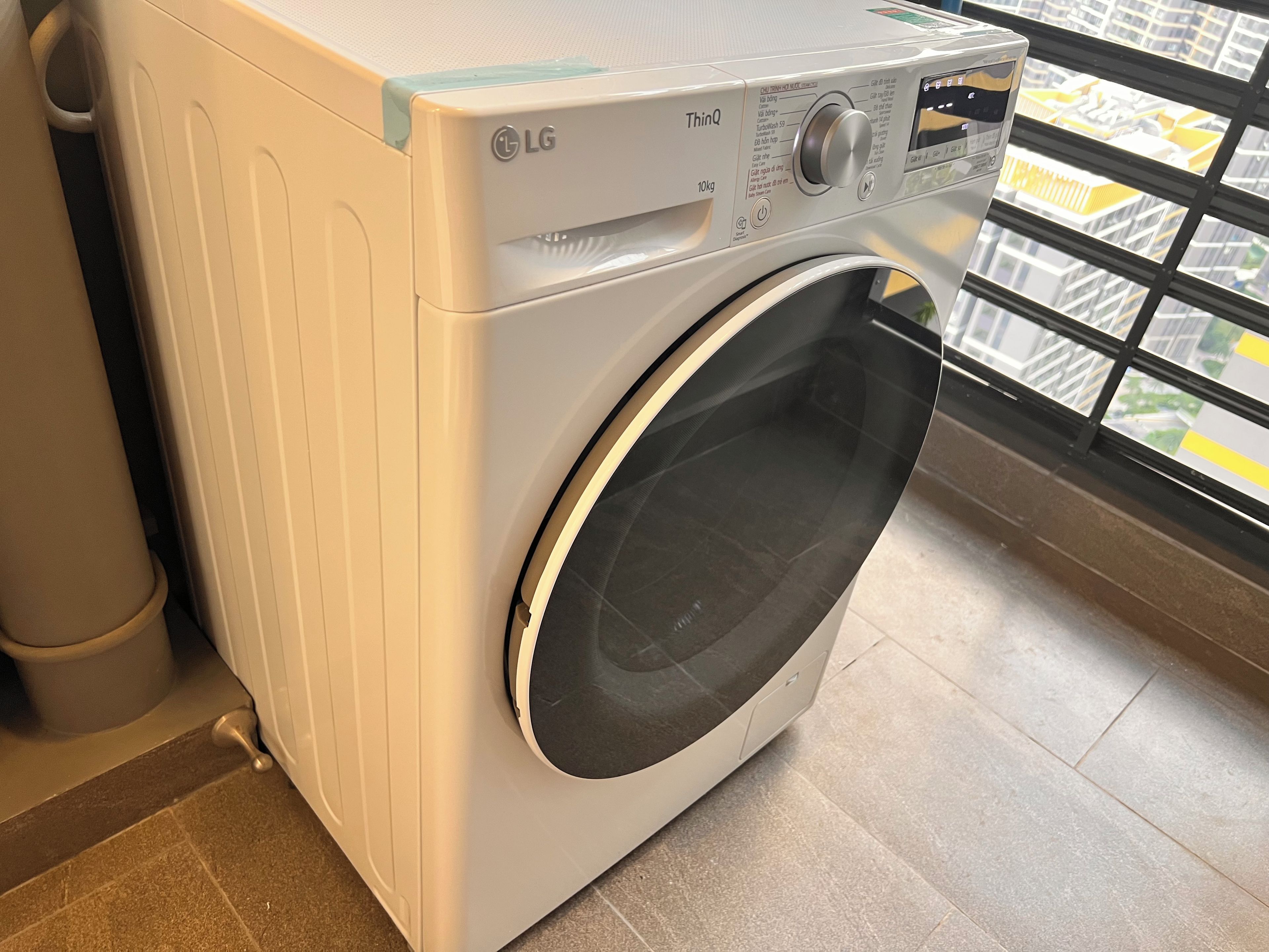 Lavadora LG ThinQ de carga frontal en una cocina moderna con vista a un paisaje urbano a través de una ventana.