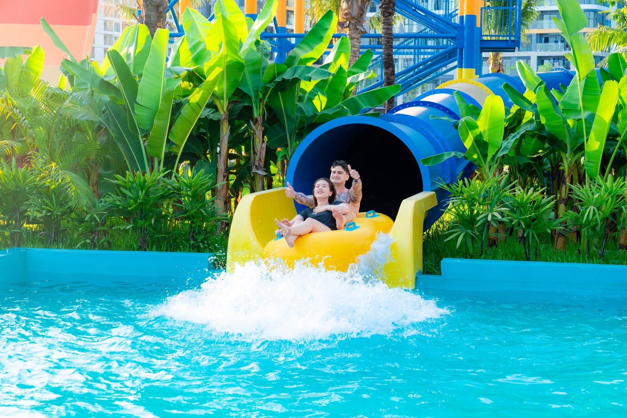 A water slide with two people at the end, surrounded by lush greenery and a blue pool.