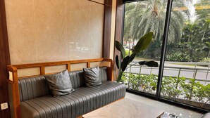 A modern interior with a leather sofa, a wooden table, and a large window offering a view of greenery and a road.