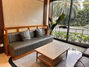 A modern interior with a leather sofa, a wooden table, and a large window offering a view of greenery and a road.