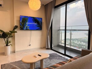 A modern living room with a large TV, a potted plant, a sofa, and a view of a cityscape through a large window.