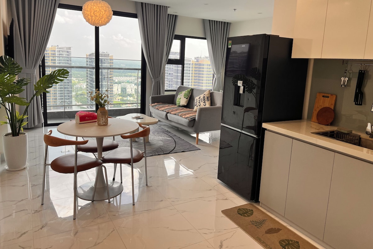 A modern living room with a sofa, dining table, and chairs, a kitchen with a refrigerator, and a large window with a city view.