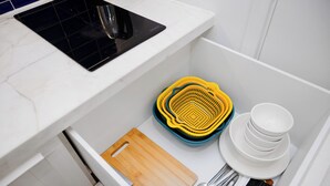 Una cocina con estufa negra, cajón para tabla de cortar de madera, un juego de coladores amarillo y azul, y vajilla blanca.