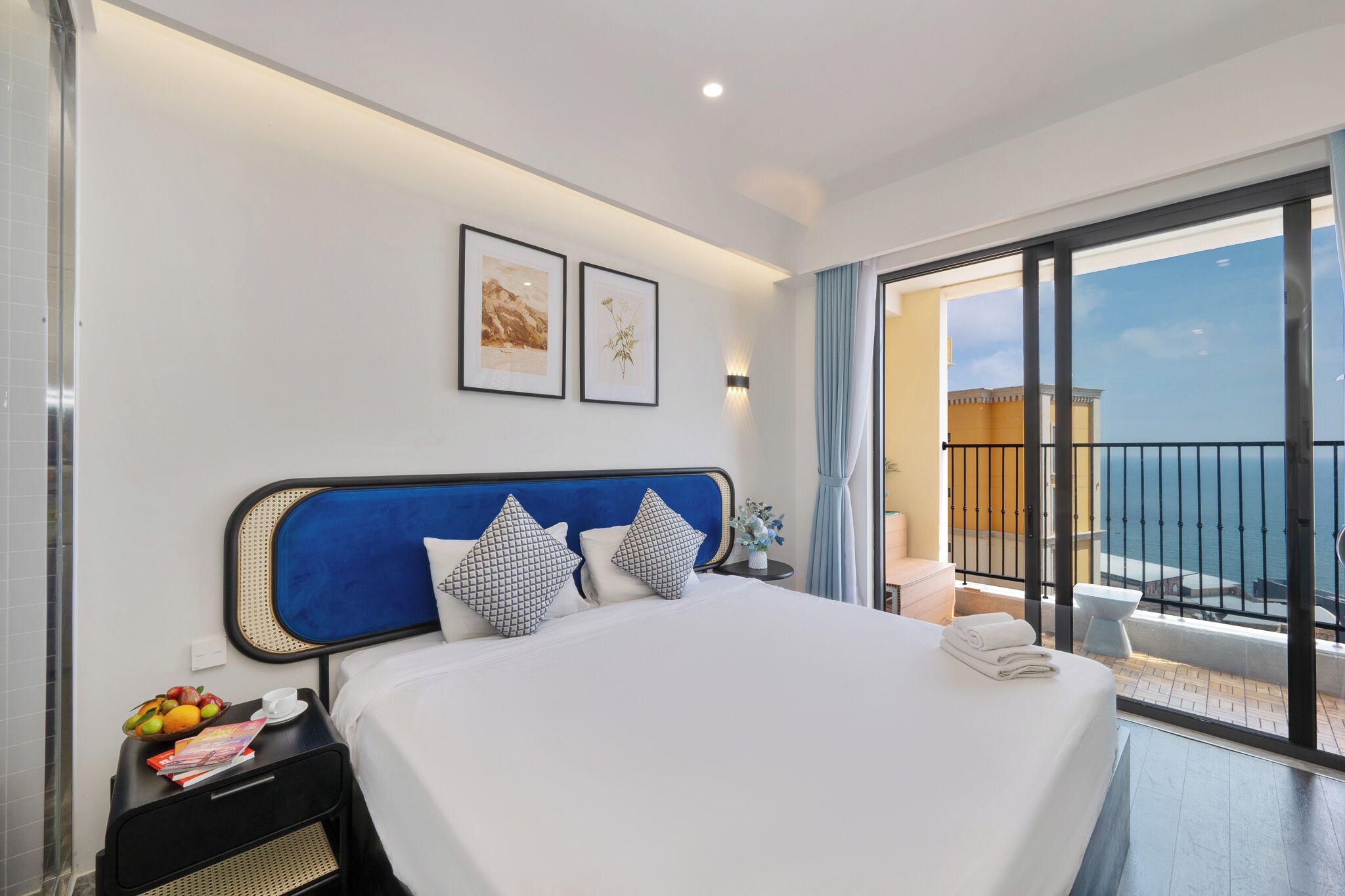 A hotel room with a large bed, a nightstand with a fruit bowl, a framed picture, and a view of the sea through a balcony.
