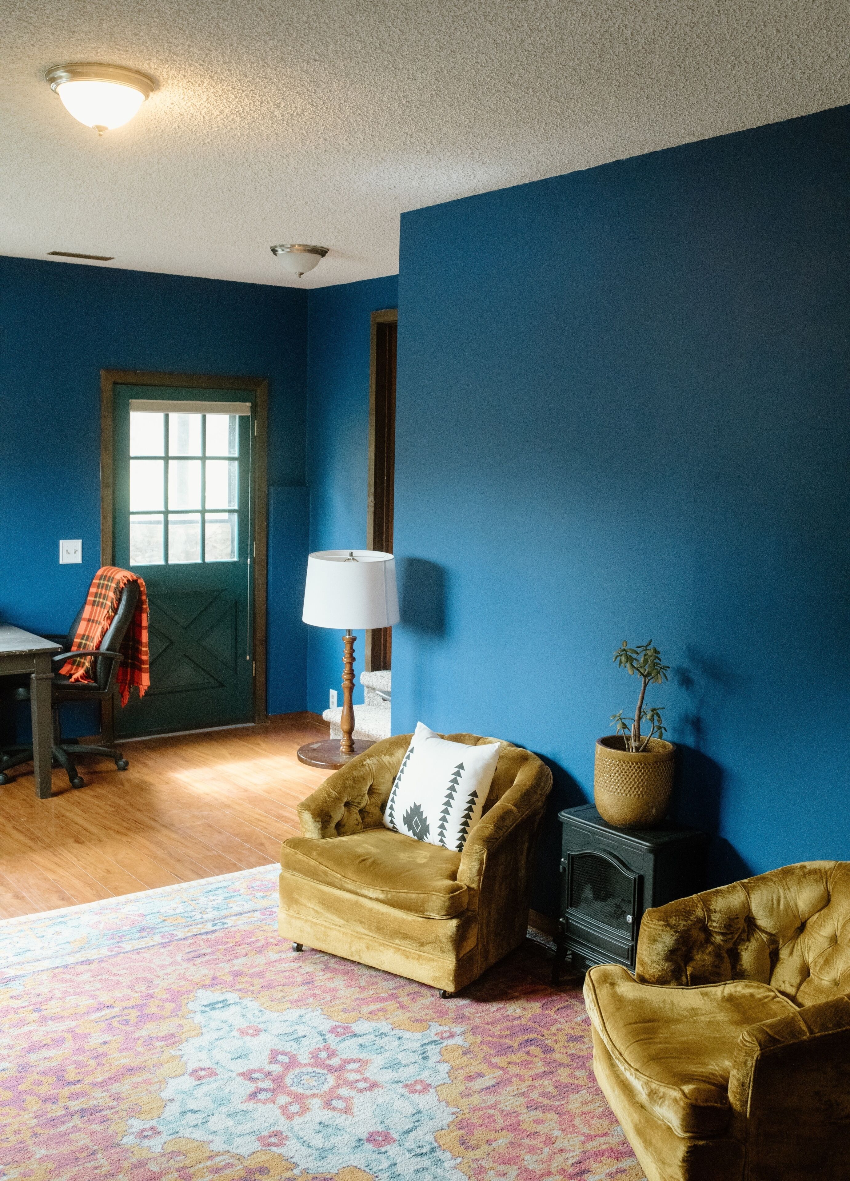 Une pièce aux murs bleus, avec un plancher en bois, un canapé, une chaise, une lampe, une plante et une porte dotée d’une fenêtre.