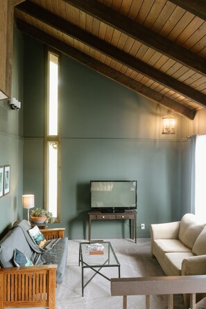 A living room with a television on a stand, a sofa, a coffee table, and a window with curtains. - Northwood Lodge: 4BR, Sleeps 10, 1 Mile to U of I (Moscow)