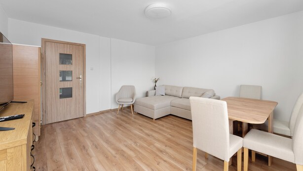 Una sala de estar moderna y minimalista, con piso de madera, un sofá color beige, una mesa de comedor con cuatro sillas y una puerta de madera con paneles de vidrio.
