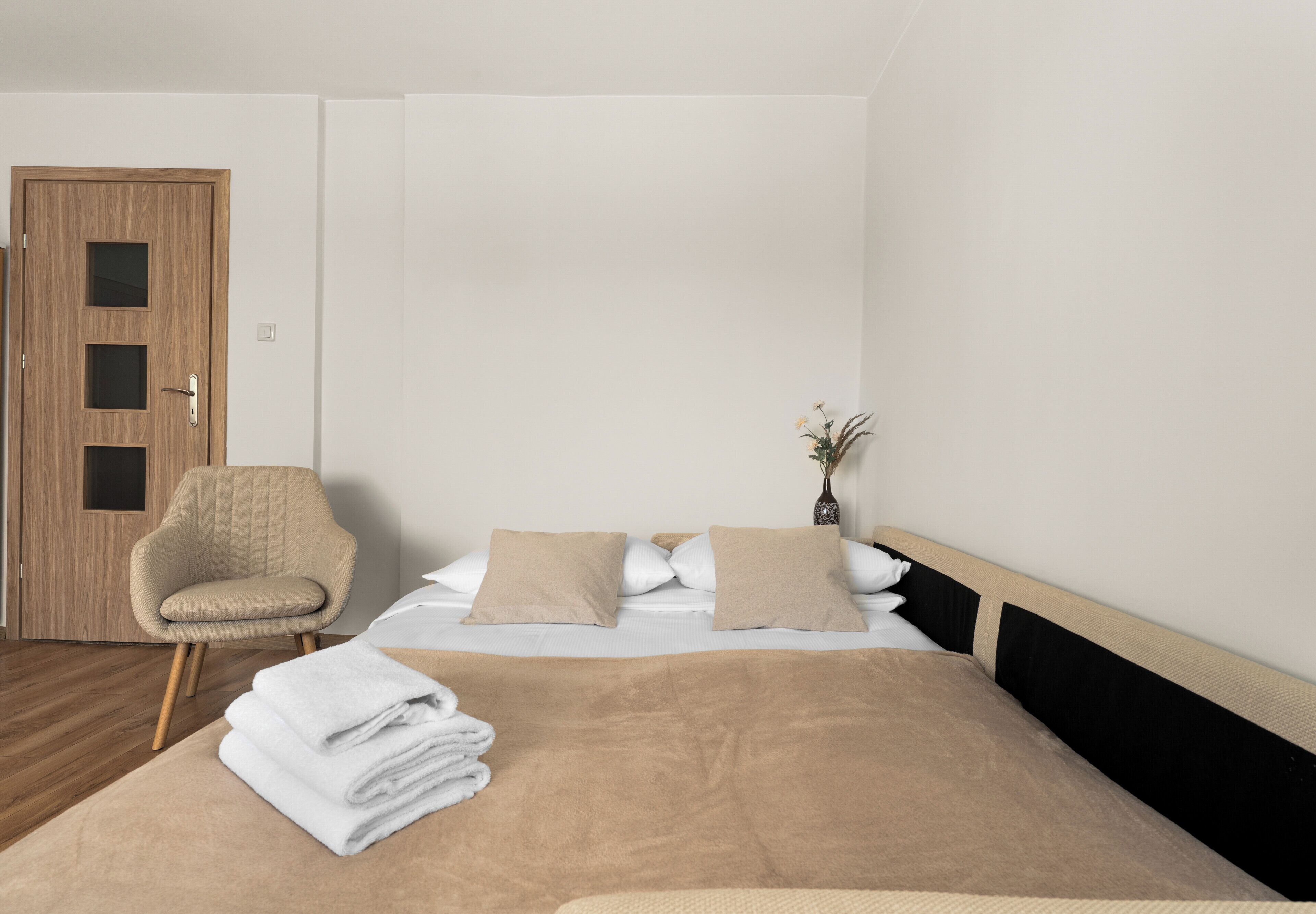 Quarto com cama, uma cadeira, porta e um vaso com flores.