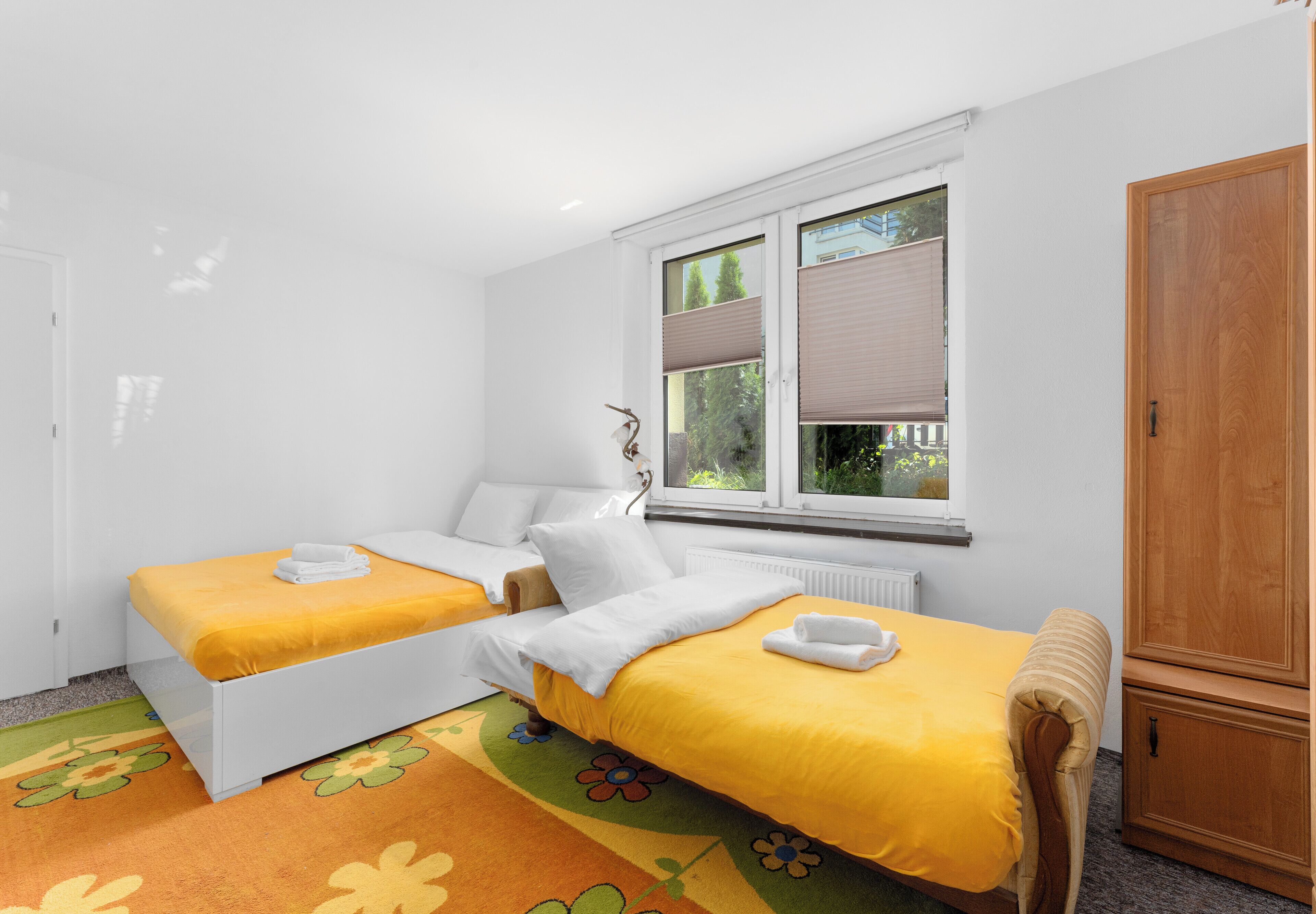 A room with two beds, a radiator, a window with blinds, and a wooden wardrobe.