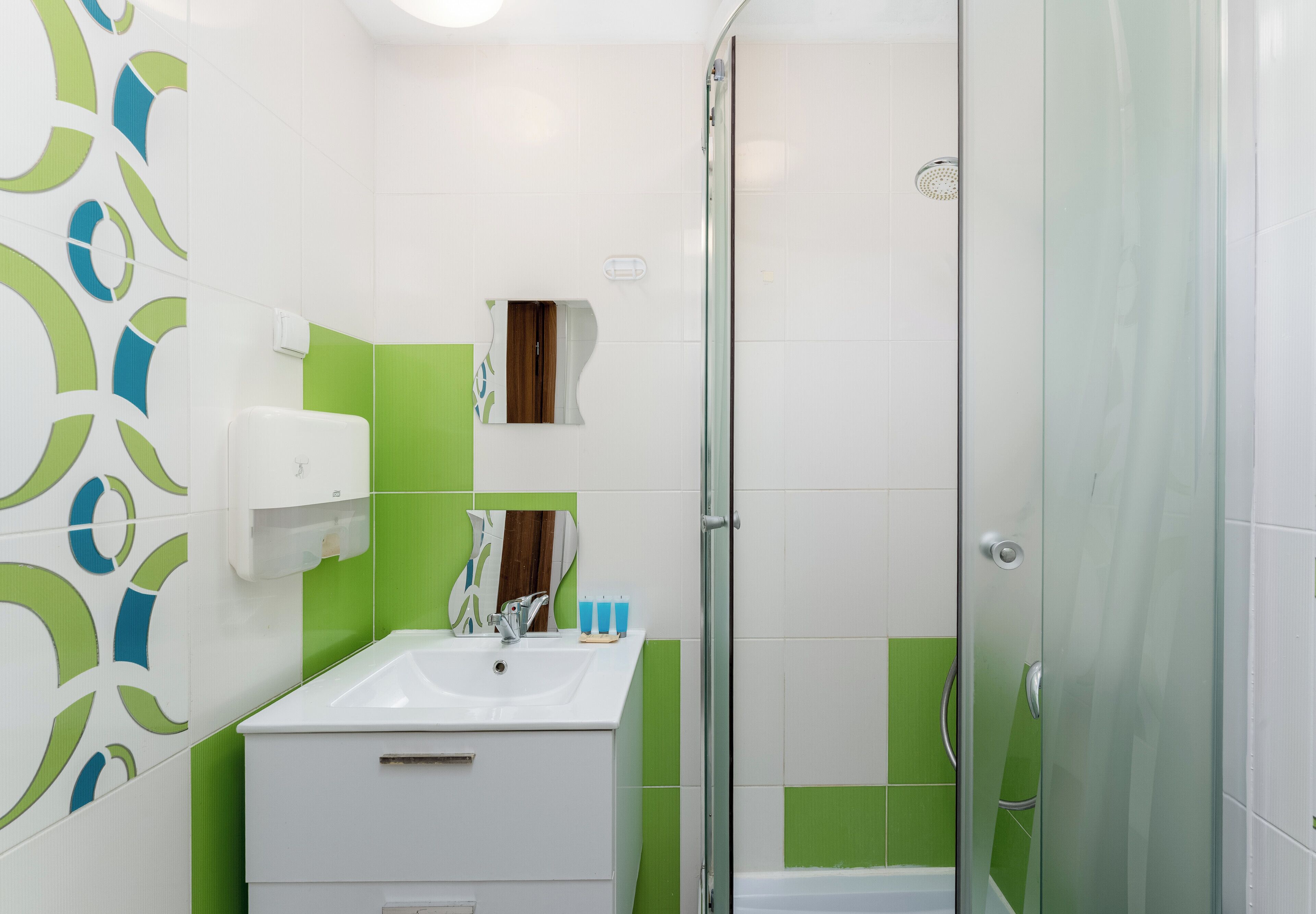 A bathroom with a white sink, green and white tiles, a glass shower enclosure, and a mirror with a wooden frame.