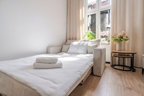 A neatly made bed with white linens and a folded towel, a bedside table with a vase of flowers, and a window with curtains. - Słowackiego Chic Apartment (Katowice)