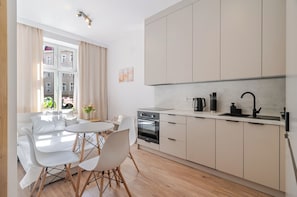 A modern kitchen with a dining area, featuring a white table, chairs, and a view of buildings through the window. - Słowackiego Chic Apartment (Katowice)