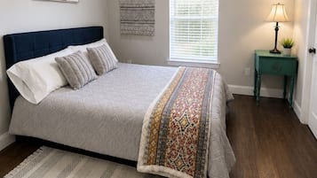 Un dormitorio con una cama, ventilador de techo, ventana con persianas, mesita de noche con lámpara y una obra de arte colgada en la pared.