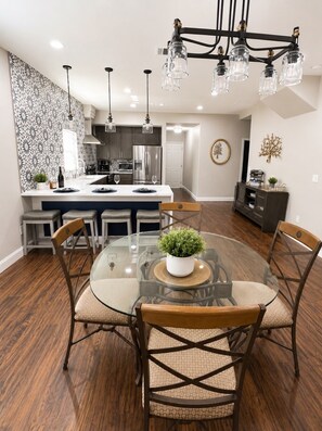 Una cocina moderna con electrodomésticos de acero inoxidable, un comedor con mesa de vidrio y sillas de madera, y un papel tapiz estampado en la pared izquierda.