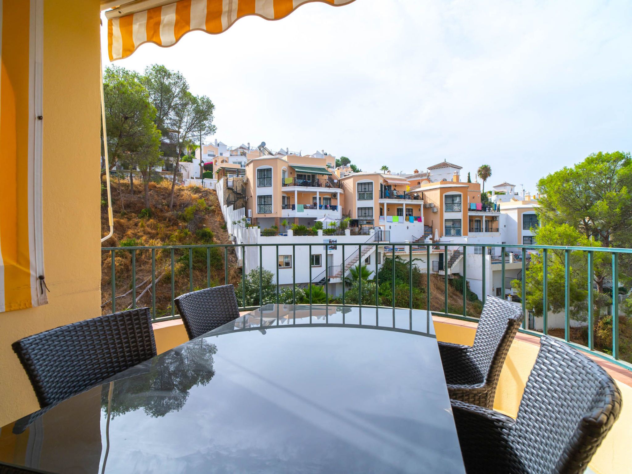 A balcony with a glass table and wicker chairs, overlooking a hillside of residential buildings.