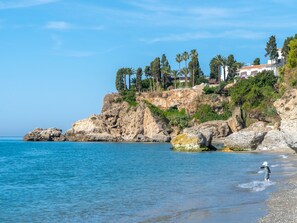 A coastal landscape with rocky cliffs, a clear blue sea, and a building with a red roof on the hillside. - Las Flores de Burriana 5 by Casasol by Interhome (Nerja)