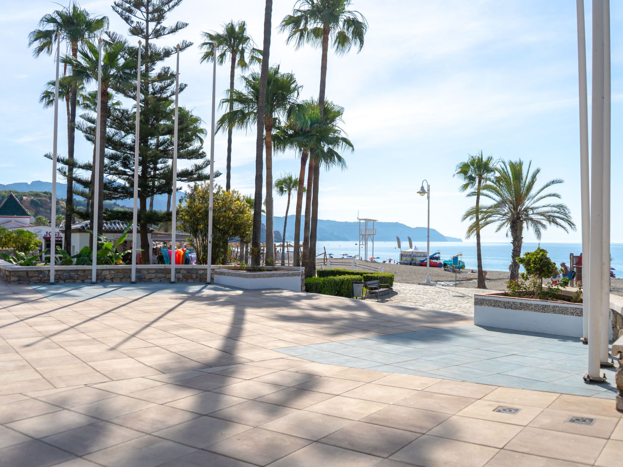 Eine gepflasterte Promenade mit Palmen, Bänken und Blick auf Strand und Meer.