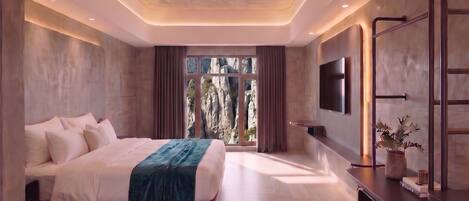 A modern bedroom with a large bed, a TV mounted on the wall, and a view of rocky terrain through the window.