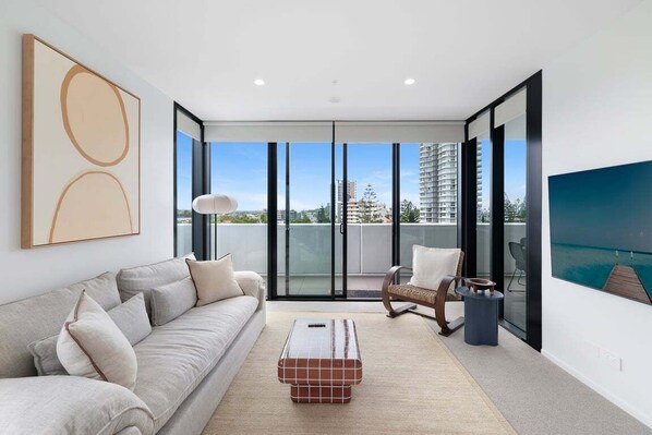 Un salon moderne avec un canapé, une table basse et un fauteuil. Une grande fenêtre offre une vue sur les immeubles et donne accès à un balcon.