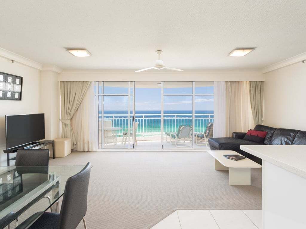 A spacious living area with a balcony overlooking the sea, a flat-screen TV, a dining table, and a sofa.