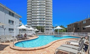 A swimming pool with lounge chairs and umbrellas, surrounded by a white fence, with a tall building in the background. - 2nd Avenue Apartments - 13B (2B/2B) (Gold Coast)