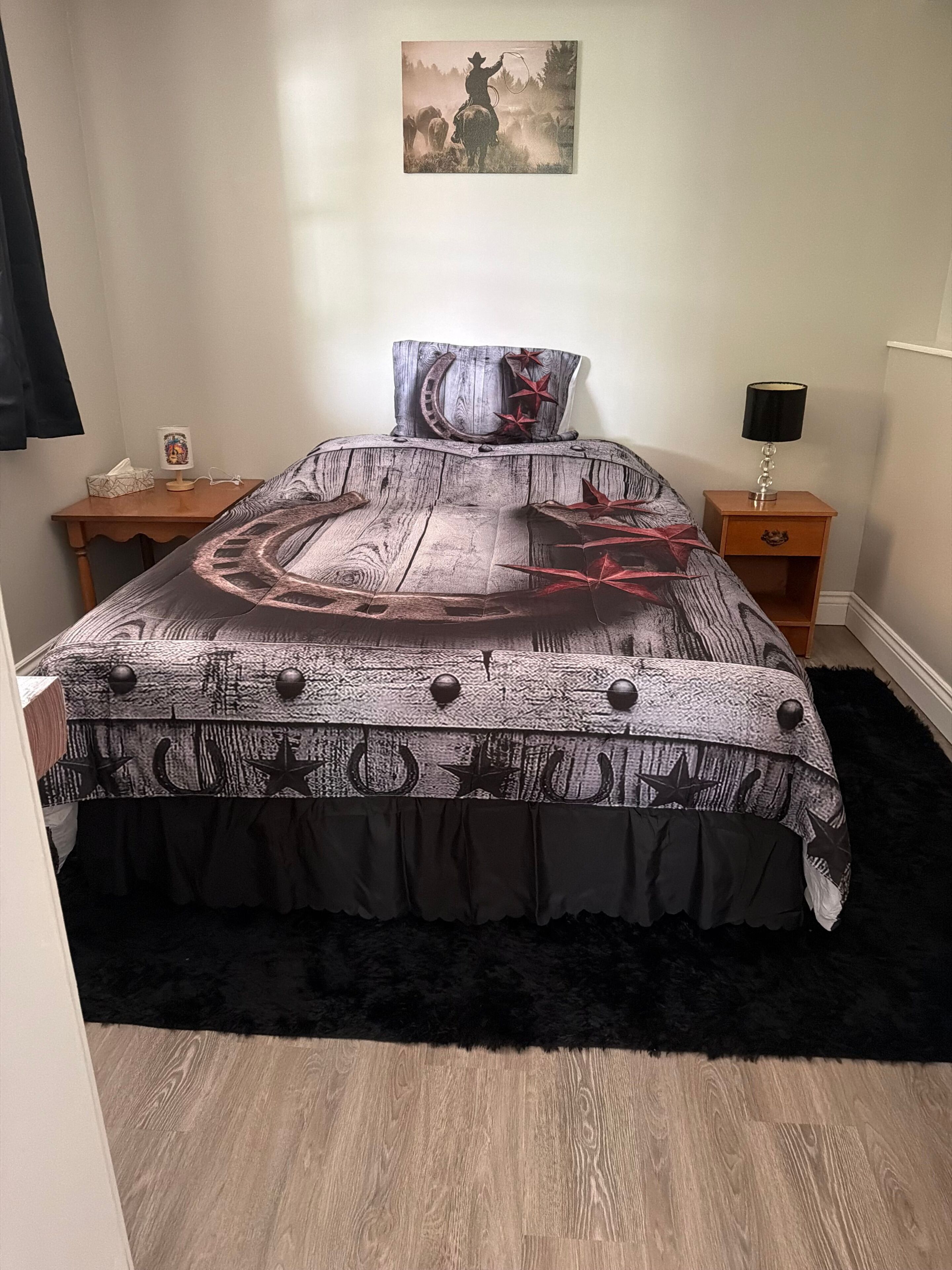 A bedroom with a bed, a nightstand, a lamp, a wall-mounted painting, and a wooden floor.