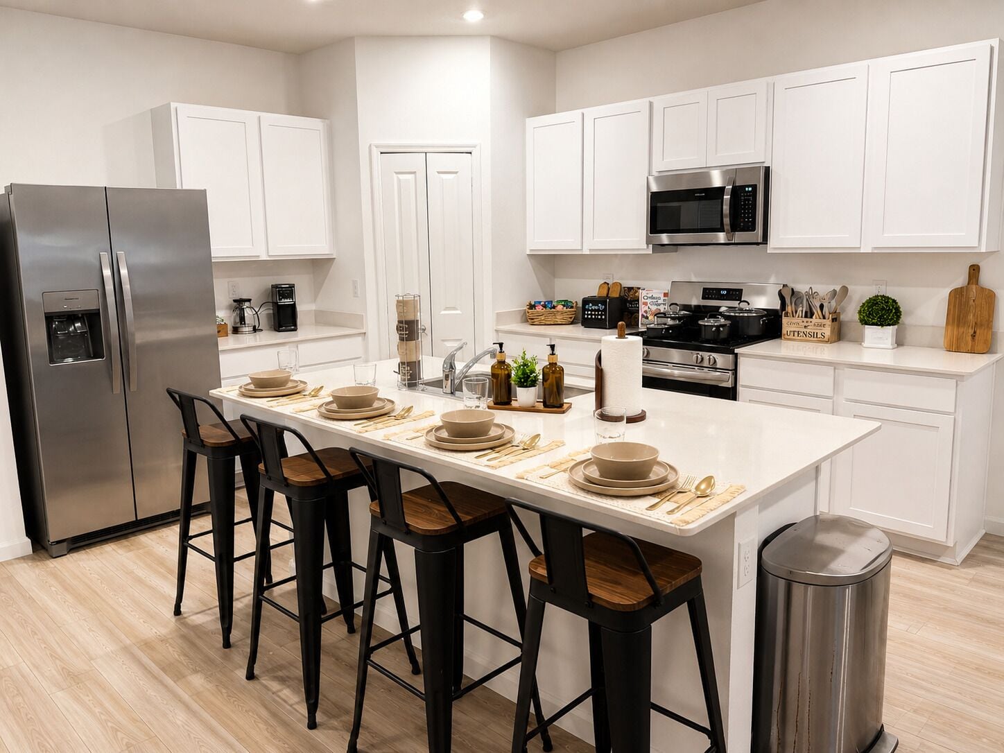 Una cocina moderna con armarios blancos, un refrigerador de acero inoxidable, un microondas y una estufa. Hay taburetes de bar y una mesa de comedor con platos y cubiertos.
