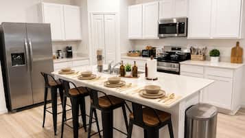 Una cocina moderna con armarios blancos, un refrigerador de acero inoxidable, un microondas y una estufa. Hay taburetes de bar y una mesa de comedor con platos y cubiertos.