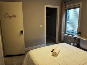 Ein Hotelzimmer mit einem Bett, einer mit 'Coyote' beschrifteten Tür, einem Fenster mit Jalousien und einem kleinen Tisch mit einer Pflanze.