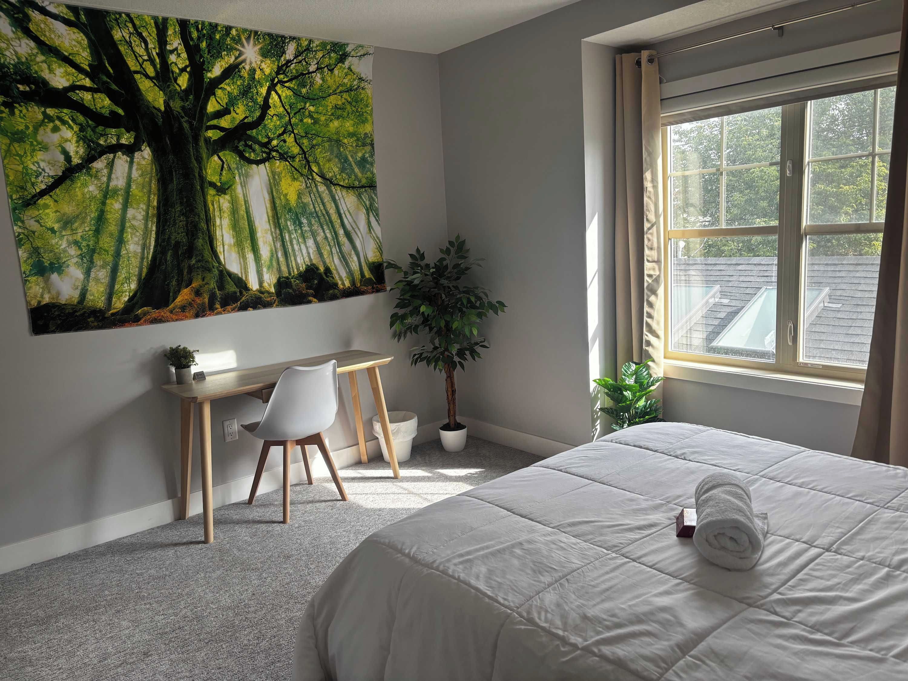 A bedroom with a bed, a desk, a chair, a large painting of a forest, and a window with a view of the outside.