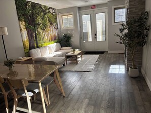 A modern living room with a sectional sofa, a dining table with chairs, a coffee table, and a large forest mural on the wall. - Fox - Private Bedroom near Downtown and 17th Av (Calgary)