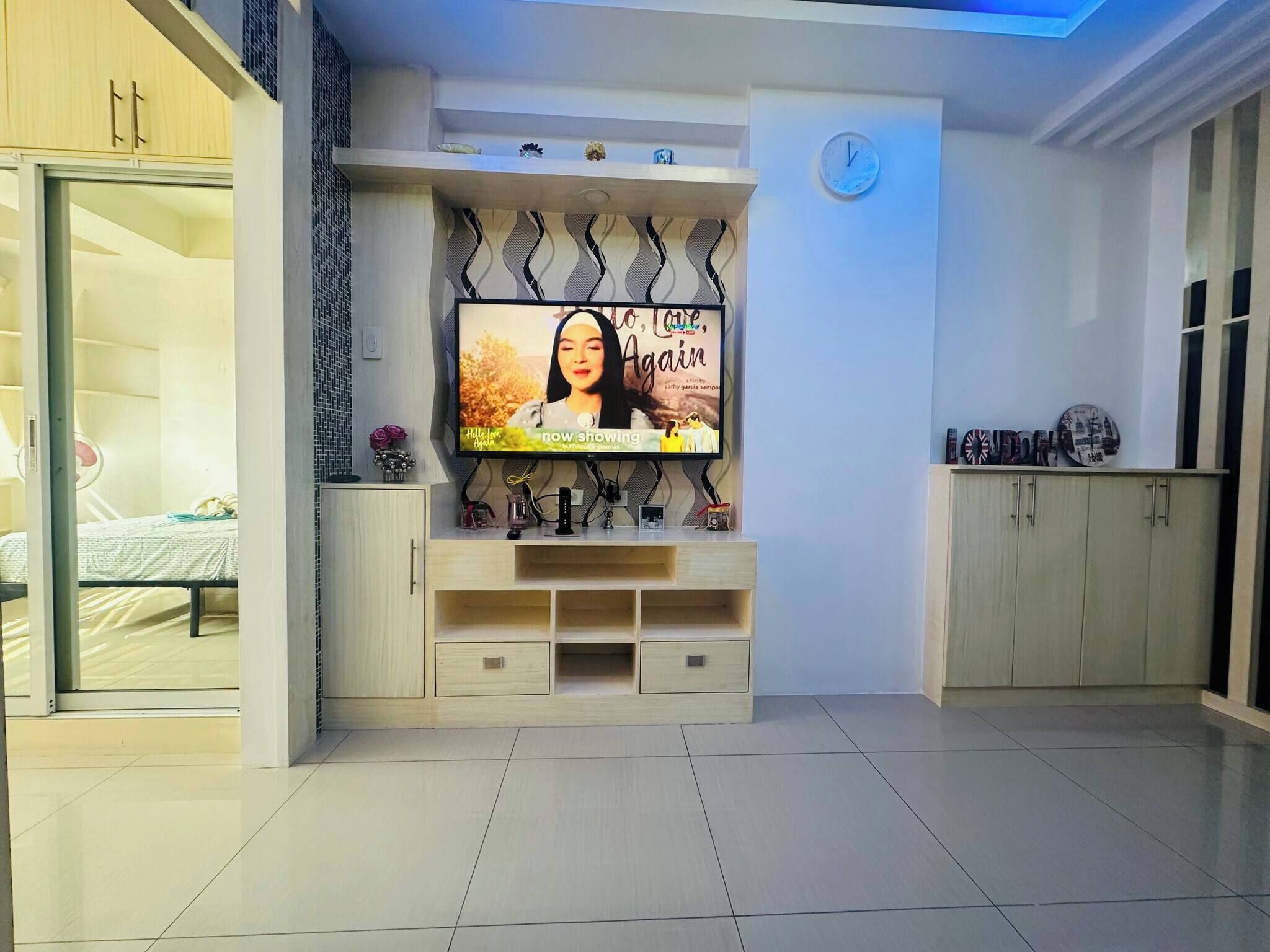 Una sala de estar moderna con un televisor de pantalla plana montado en la pared, una cama con cabecera blanca y un aparador de madera con objetos decorativos.