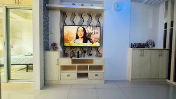 Una sala de estar moderna con un televisor de pantalla plana montado en la pared, una cama con cabecera blanca y un aparador de madera con objetos decorativos.