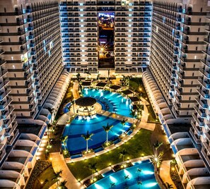 Ein mehrstöckiges Hotel mit einem zentral gelegenen Pool, umgeben von Liegebereichen und Palmen.