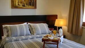 Habitación de hotel con cama, una mesita de noche con cosas para el desayuno, una lámpara y un cuadro en la pared.