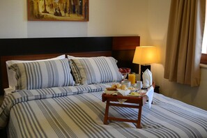 Habitación de hotel con cama, una mesita de noche con cosas para el desayuno, una lámpara y un cuadro en la pared.