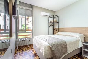 A bedroom with a bed, a window, an air conditioning unit, and a wooden floor.