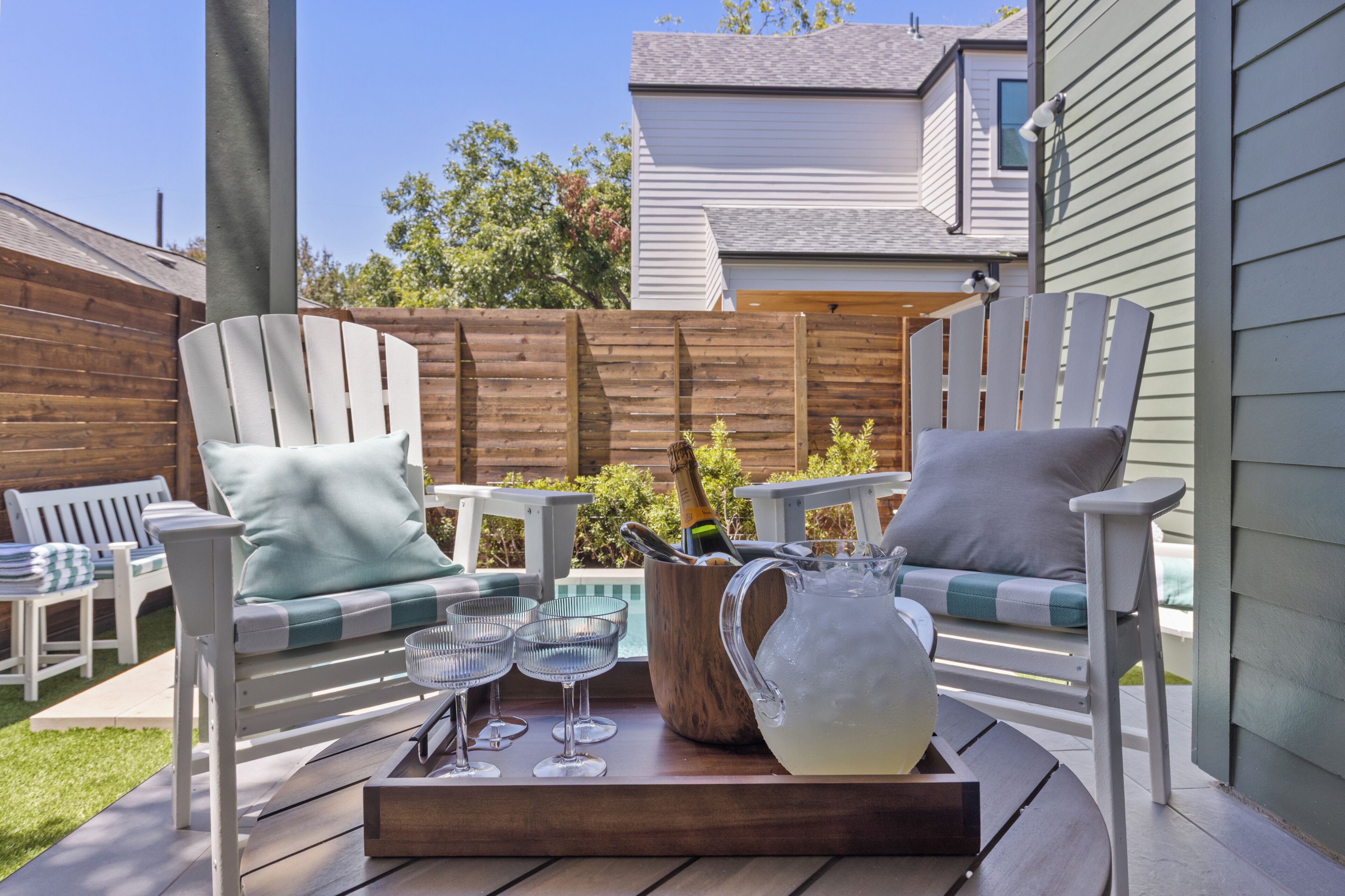 An outdoor patio with Adirondack chairs, a wooden table with a pitcher and glasses, and a pool in the background.