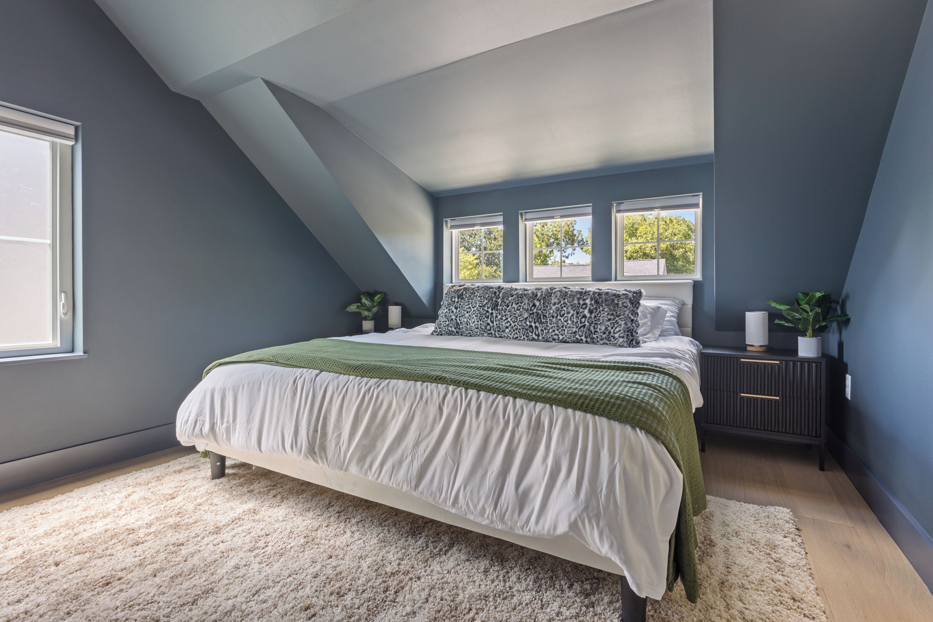 A bedroom with a sloped ceiling, a large bed with a green and white bedspread, two windows, and a nightstand with a plant.