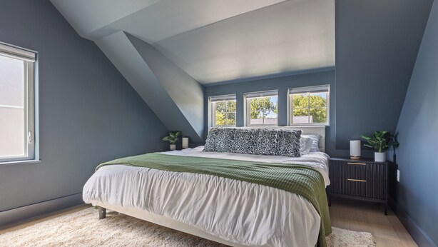 A bedroom with a sloped ceiling, a large bed with a green and white bedspread, two windows, and a nightstand with a plant. - Beautiful 4-bedroom - Pool, Patio, Grill - Close to UT & Rainey (Austin)