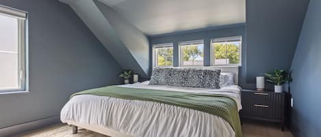 Un dormitorio con techo inclinado, una cama grande con colcha verde y blanca, dos ventanas y una mesita de noche con una planta.