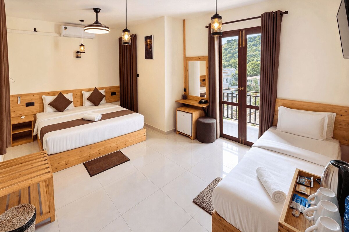 A hotel room with two beds, a desk, a small kitchenette, and a balcony with curtains.