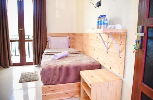 Una habitación pequeña con cama individual, cabecera de madera, un banco y una mesita. Se observa una ventana con cortinas, una puerta con manija y un estante con botellas de agua y vasos.