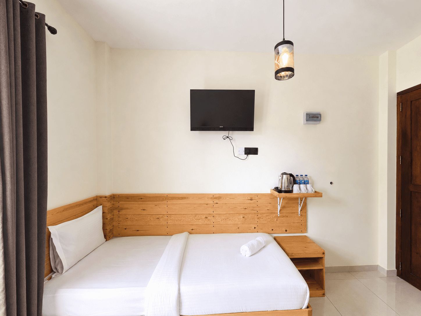 A hotel room with a wooden headboard, a flat-screen TV mounted on the wall, a bedside table with a kettle and bottled water, and a wooden door.