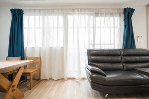 A room with a wooden dining table, chairs, a black leather sofa, and a window with sheer curtains and solid blue curtains. - SERENE | Nishikujo, 5th Floor | 100 ㎡, Sleeps 10 | Direct Access to USJ and Osaka Station (Konohana Ward)