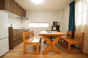 A compact kitchen with wooden cabinets, a dining table with benches, a microwave, and a window with curtains. - Aoba Residence ASAHI: 2-Minute Walk to Nishikujo Subway Station, 100 sqm, 4-Bedroom Residence Near USJ (大阪市)